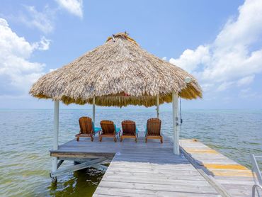 Thatched-roof hut with lounge chairs on a wooden pier over calm water.