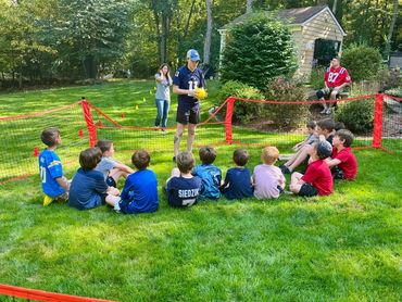 Kids sitting on grass listening to an instructor holding a yellow ball near a net.