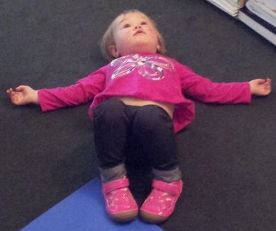 A toddler lying on the floor with arms outstretched wearing a pink top and shoes.