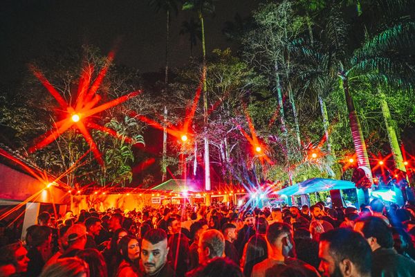 Crowded outdoor night party with vibrant red lights and tall trees.