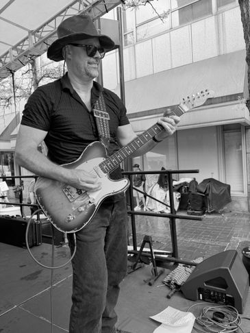 Man playing electric guitar on an outdoor stage, wearing a hat and sunglasses.