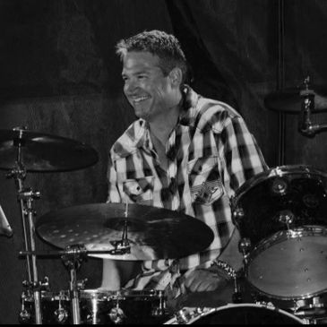 Smiling man playing drums in a black-and-white photo.
