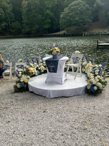 Elegant lakeside wedding setup with floral arrangements and white chairs.