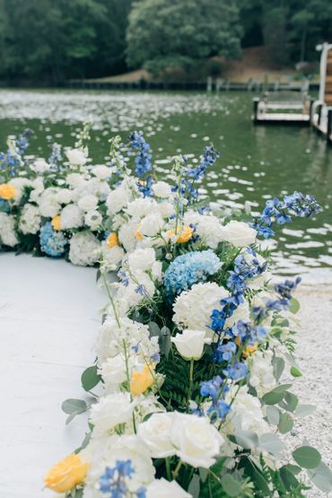 Elegant floral arrangement by a lakeside dock, featuring white, blue, and yellow flowers.