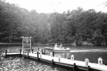 A couple on a boat near a decorated dock on a lake surrounded by trees.