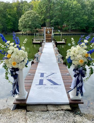 A decorated dock with flowers and a white aisle runner for a wedding ceremony by the water.