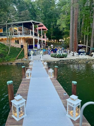 A decorated dock leading to a lakeside gathering with string lights and floral arrangements.