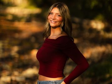 A smiling woman in a maroon top and jeans poses outdoors in warm sunlight.