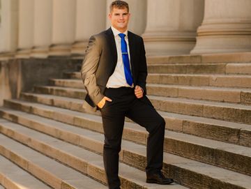 A young man in a suit stands confidently on stone steps outdoors.