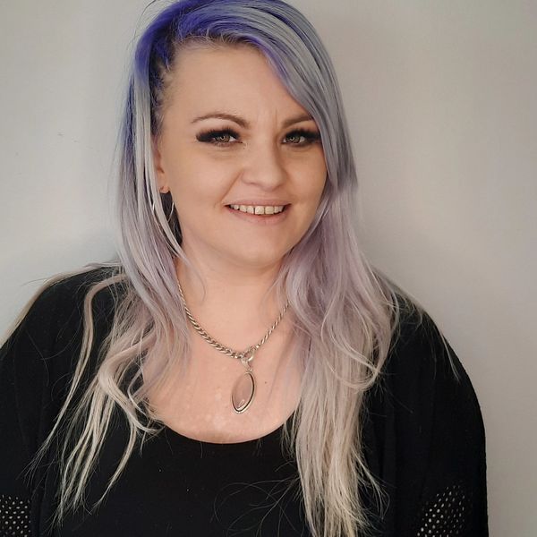 Smiling woman with long lavender hair and a silver necklace against a plain background.