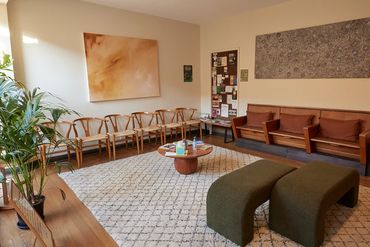 Cozy waiting room with wooden chairs, green stools, plants, and abstract art on beige walls.