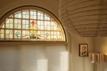 Arched stained glass window with blinds, next to a lamp and abstract wall art.