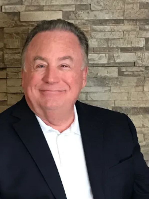 Smiling man in a dark blazer and white shirt poses against a stone wall.