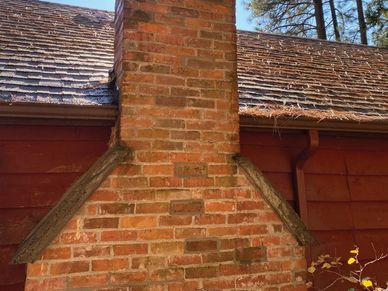 Sunlit brick chimney on a rustic house roof surrounded by tall pine trees.