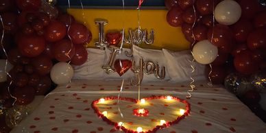 A romantic bed covered with red rose petals and heart shaped tea lights and red balloon garlands.