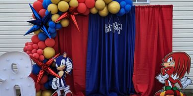 Blue and red curtain backdrop draped by a balloon garland and Sonic and Knuckles character cut-outs.