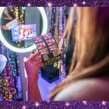 Woman standing in front of a photo booth, holding a photo booth picture looking at it with a smile.