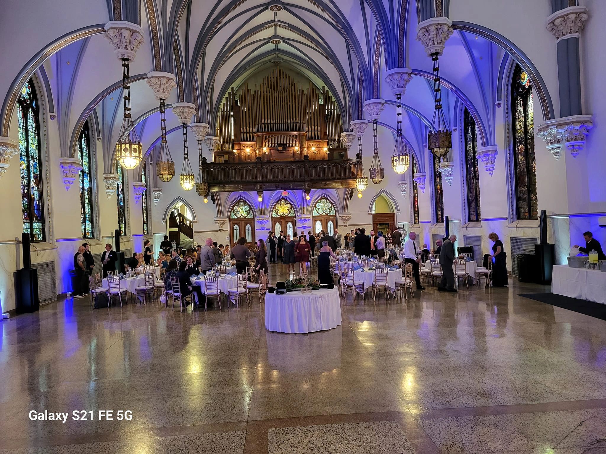 Guests mingle in a grand, gothic-style hall with blue lighting and stained glass windows.