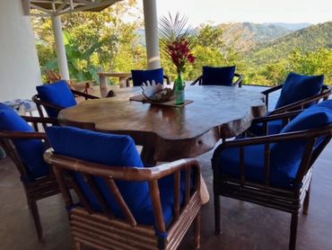 Rustic wooden table with blue cushioned chairs on a patio overlooking mountains.