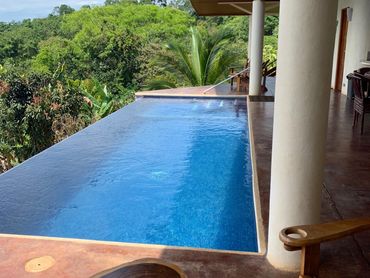 Infinity pool overlooking lush green forest from a patio with wooden furniture.