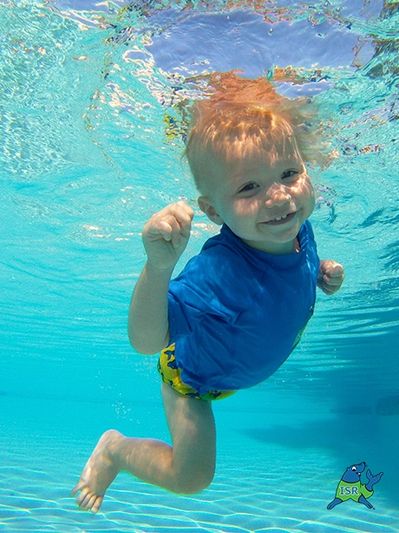 Smiling toddler swimming underwater in a pool wearing a blue shirt.