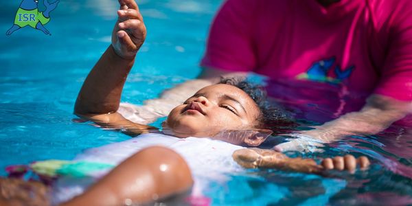 A young child learning to float in a swimming pool with an instructor's support.