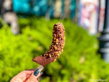 Hand holding chocolate-covered frozen banana with nuts outdoors.