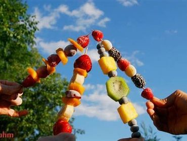 Four hands holding colorful fruit skewers against a blue sky.