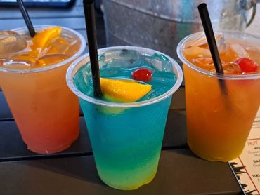 Three colorful iced cocktails in plastic cups with garnishes and straws.