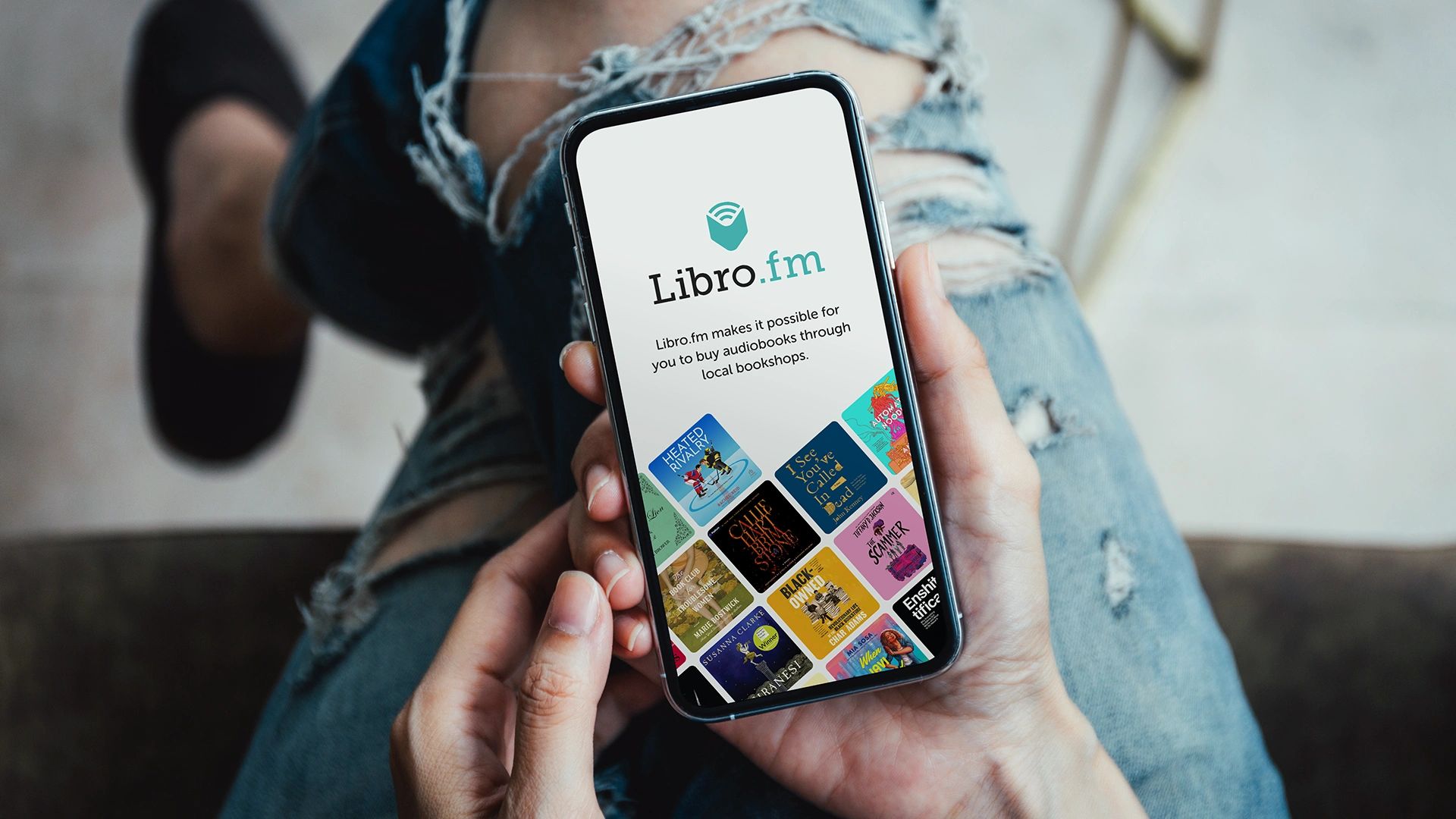 Person holding a phone displaying the Libro.fm audiobook app with ripped jeans in the background.