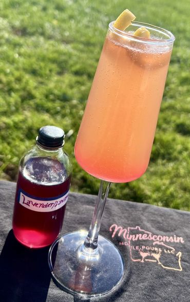 A pink cocktail with lemon garnish and a bottle of lavender syrup outdoors.