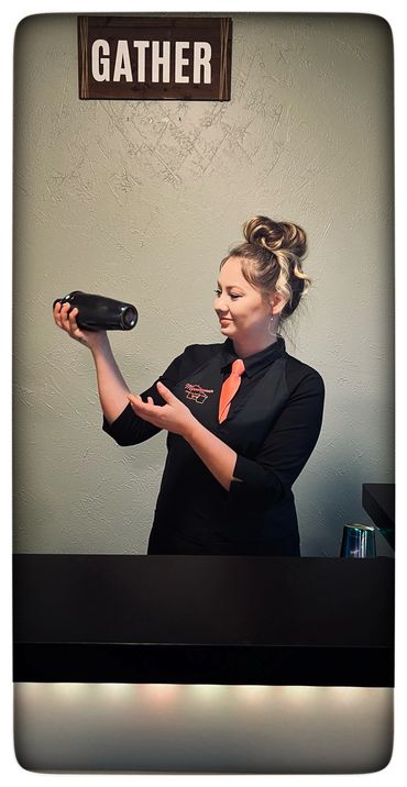 Woman bartending with a shaker under a 'GATHER' sign.