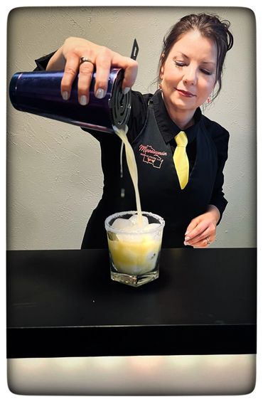 Woman pouring a yellow cocktail from a shaker into a glass with ice and a salted rim.
