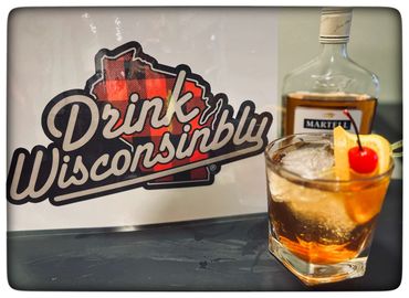 A classic cocktail with a cherry and orange garnish beside a "Drink Wisconsinbly" sign.
