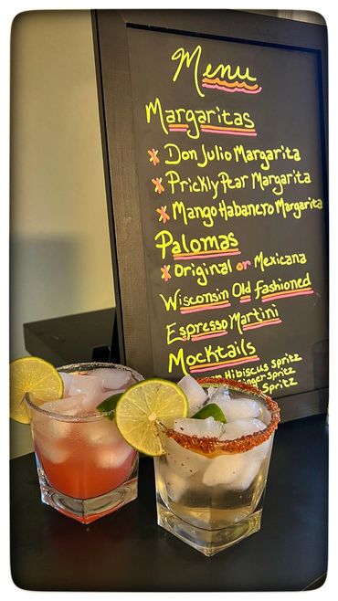 Two margaritas with lime garnishes in front of a colorful cocktail menu.