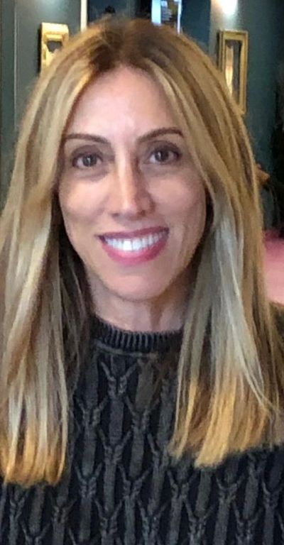 Smiling woman with long blonde hair wearing a dark patterned sweater indoors.