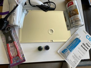 Crafting supplies including a hair dryer, mirror, silicone sealant, metallic spray paint, and rivet tool.