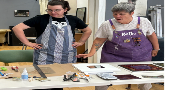 Two women in aprons discuss art pieces on a table in a studio.