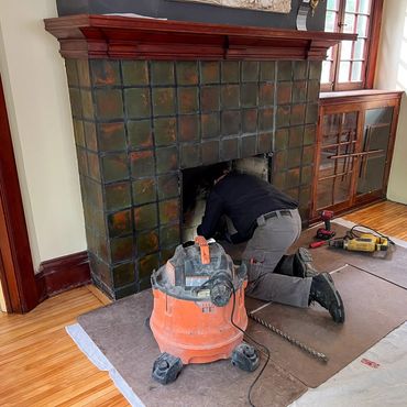 A man repairs a tiled fireplace with tools and a vacuum nearby.