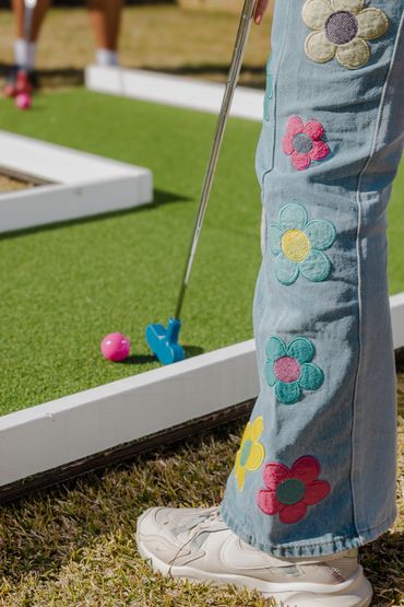 Person wearing flower-embroidered jeans playing mini golf with a pink ball.