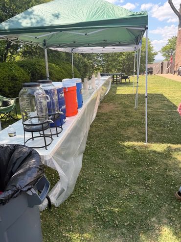 Outdoor refreshment station with coolers and water dispensers under a green canopy.