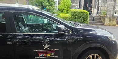 Black car with security training company logo parked by greenery.