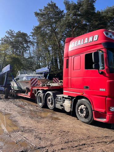 Red truck carrying a boat on a trailer in a wooded area.