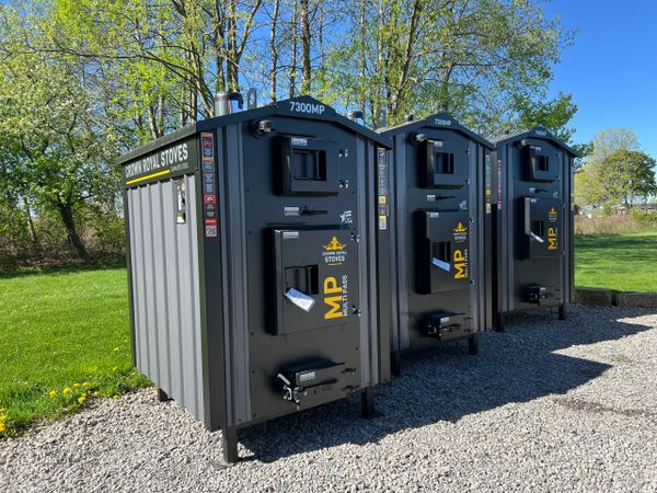 Three large black metal outdoor stoves lined up on gravel with trees in background.