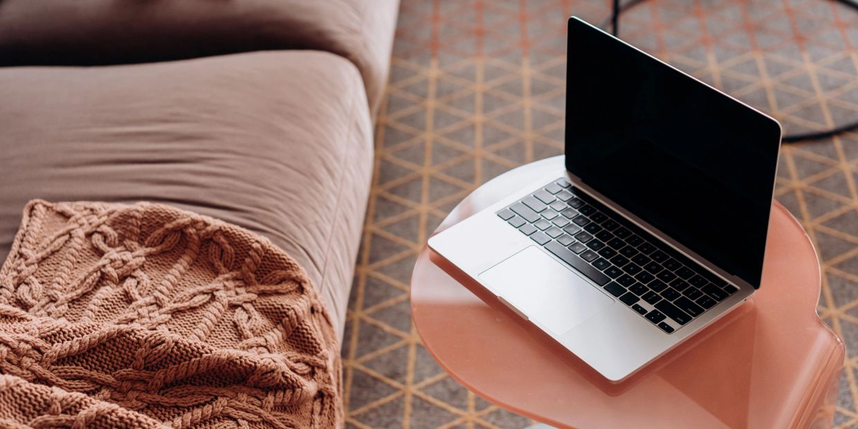 Laptop on a round table next to a brown sofa with a knitted blanket.