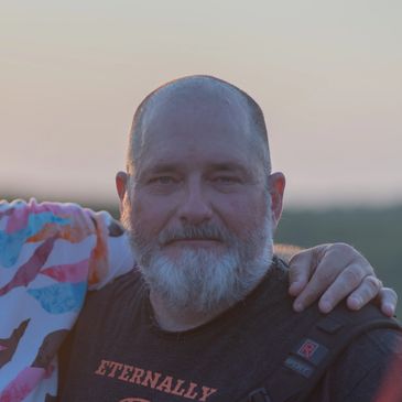 Smiling bearded man outdoors at sunset with an arm on his shoulder.