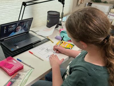 A girl with auburn hair writes with a black marker. She sits in front of a laptop on a Zoom call.
