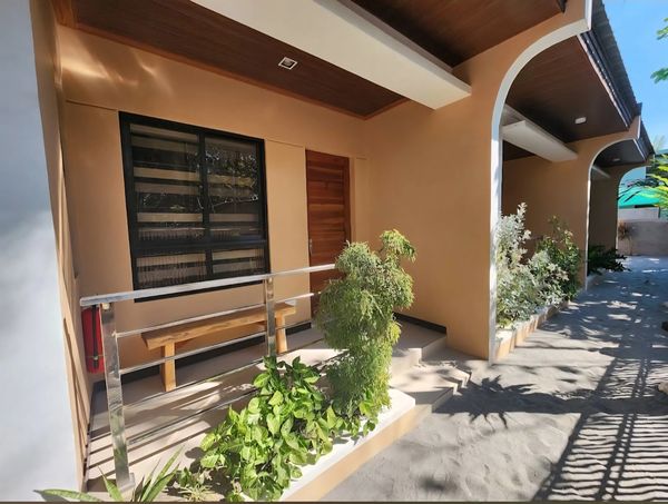 Sunlit porch with plants and bench outside a beige building.