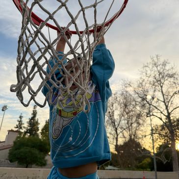 Child hanging on basketball hoop outdoors at sunset.