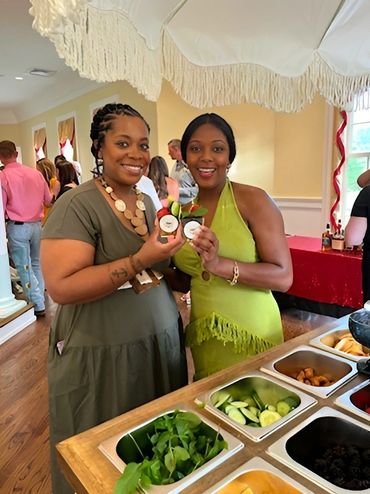 Two women smiling and holding cocktails at a social gathering with a garnish bar.